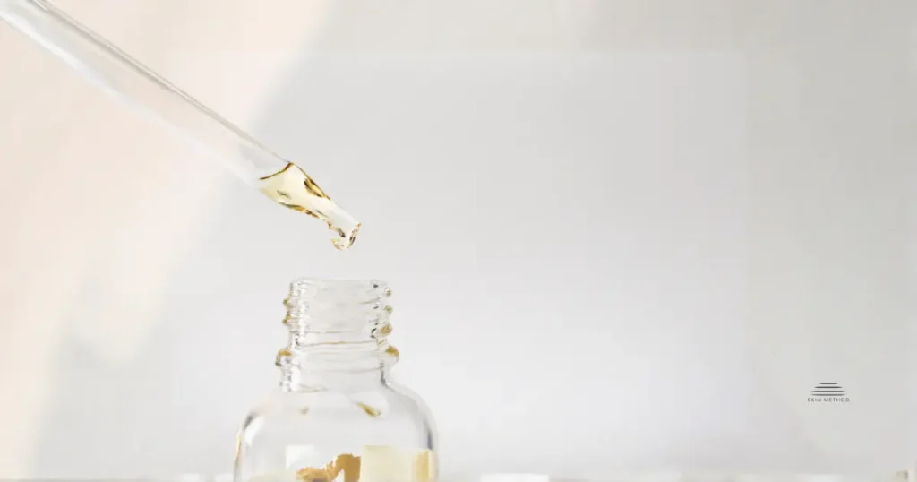 A glass dropper with pale serum suspended above an open, unbranded skincare bottle on a light neutral background.